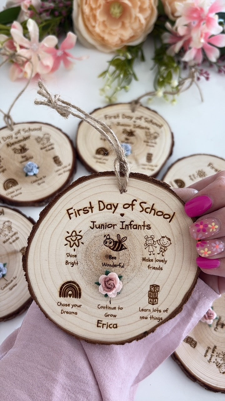 Personalized First Day of School wooden hanging ornaments displayed on a white surface with decorative flowers and fabric. A hand with pink nail polish holds the central ornament, which is engraved with the text 'First Day of School Junior Infants' and small illustrations, including a pink rose charm.