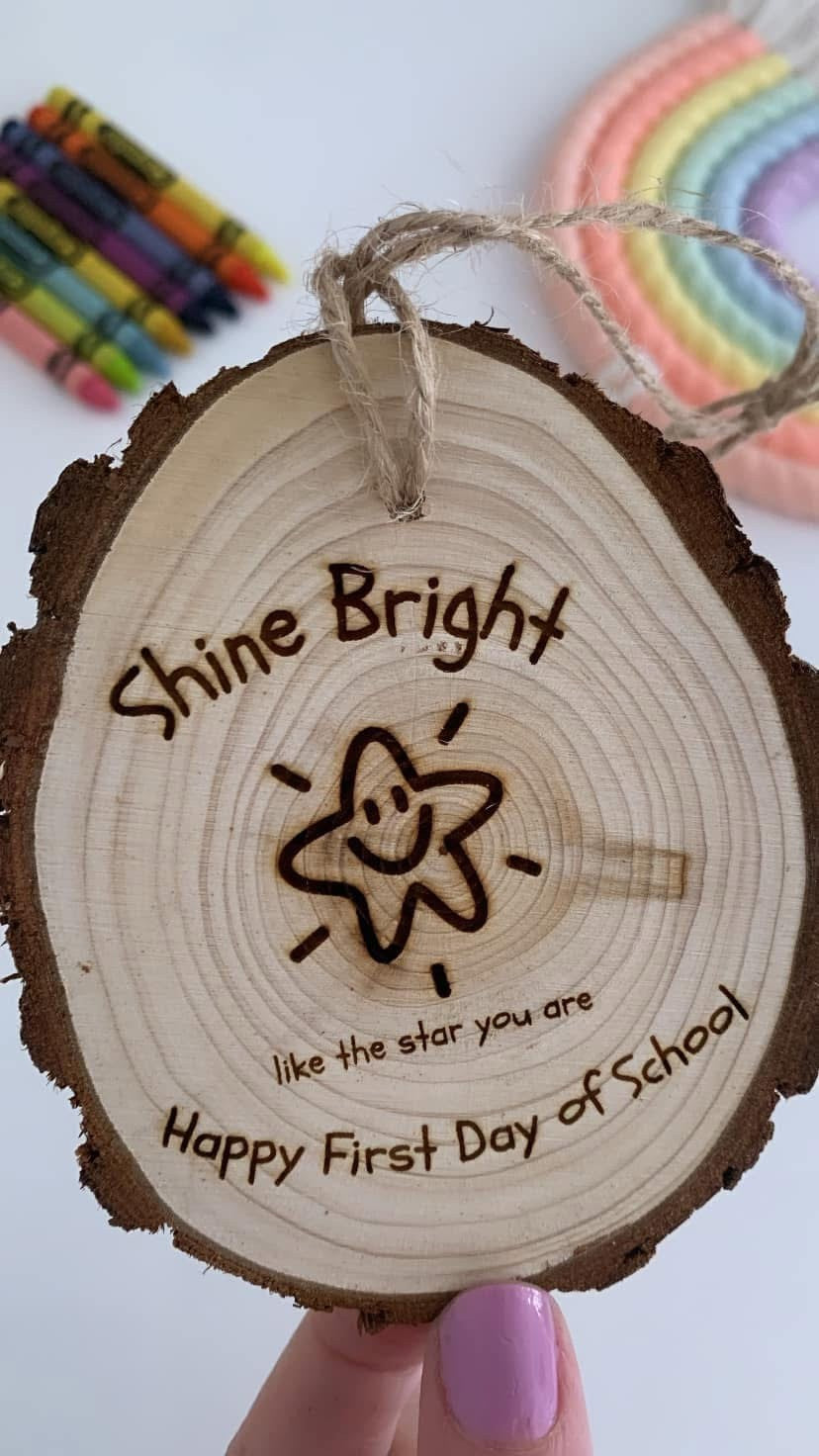 A personalized wooden slice ornament held by a hand, featuring a laser-engraved smiling star and the text 'Shine Bright like the star you are Happy First Day of School'. Crayons and a rainbow mat are blurred in the background.
