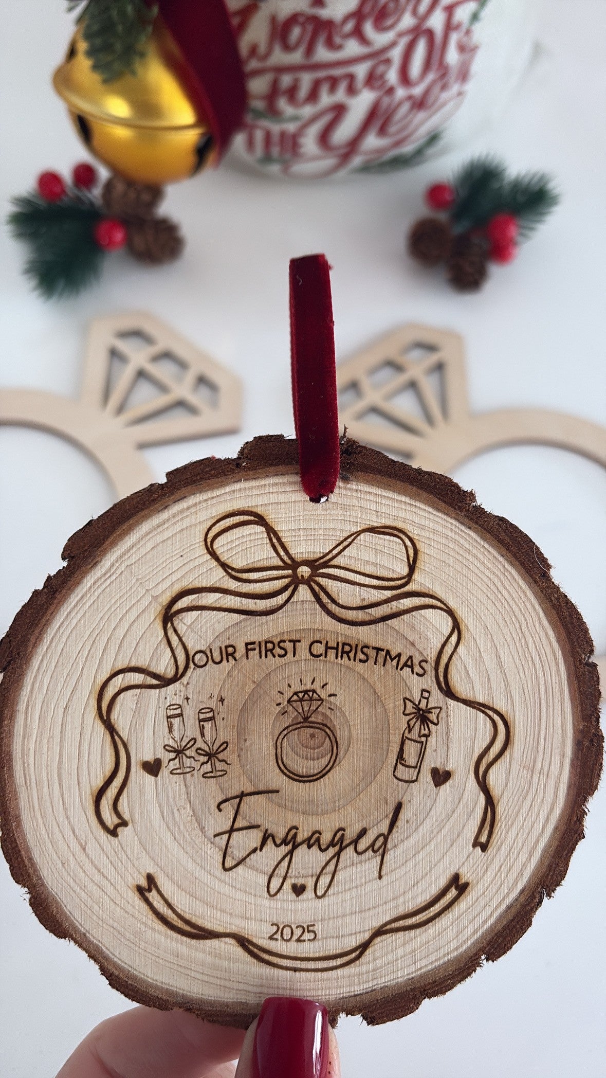 Wooden ornament with 'Our First Christmas Engaged' engraving on a white background with Christmas decorations.