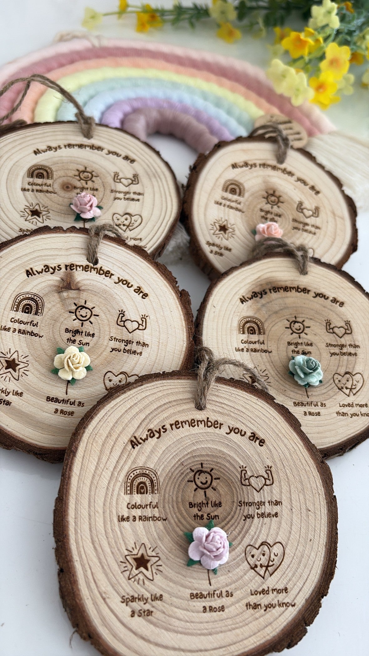 A set of engraved wooden slice ornaments with small fabric flowers and hanging loops, arranged on a light surface with a pastel rainbow decoration in the background.