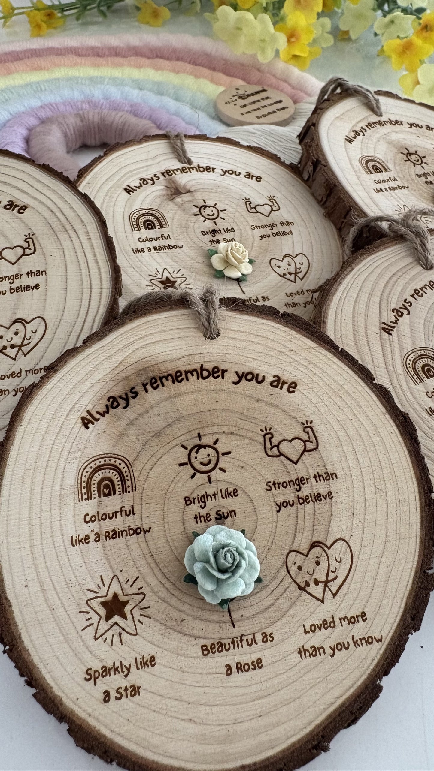 A set of personalized wooden tree-slice plaques with engraved positive affirmations, sun icons, and small hanging loops, arranged on a light surface with a rainbow mat and yellow flowers in the background.