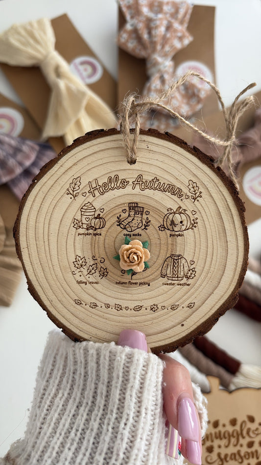 A round wooden slice hanging ornament with engraved autumn-themed illustrations and the words 'Hello Autumn' at the top, held by a hand wearing a white knit glove. The ornament has a small wooden flower in the center and twine loops for hanging.