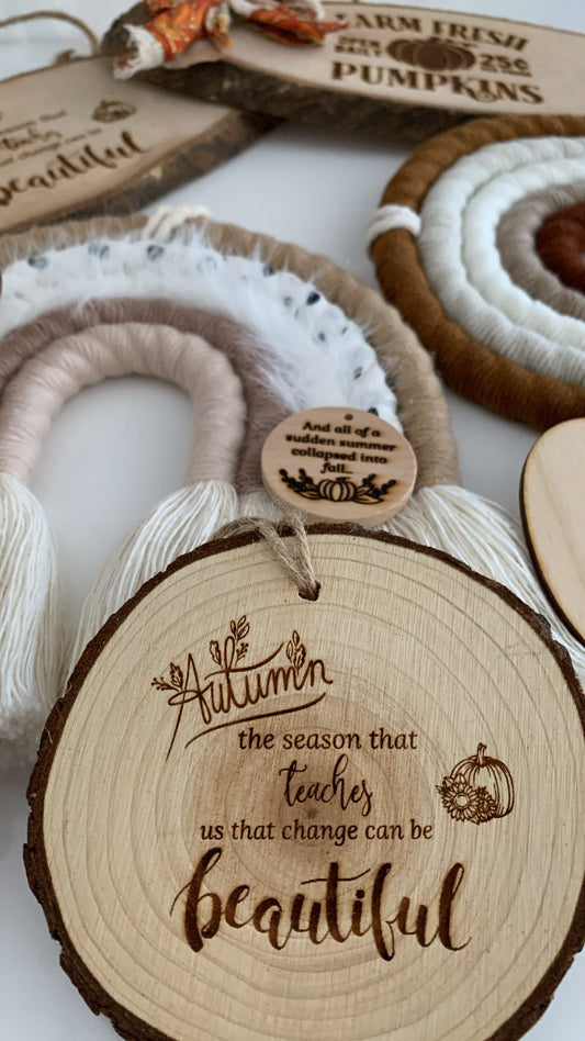 A close-up of autumn-themed wooden decor, with a round natural wood slice engraved with the phrase 'Autumn the season that teaches us that change can be beautiful' and a small hanging tag. Other visible items include woven fringe pieces and a wooden sign reading 'Farm Fresh Pumpkins'.