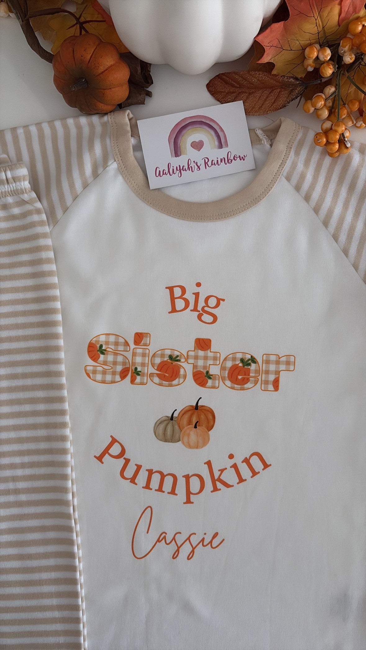 A white raglan baby shirt with beige trim, featuring a personalized graphic that reads 'Big Sister Pumpkin Cassie' in orange lettering with small pumpkin illustrations. The shirt is styled with a striped blanket, decorative pumpkins, and autumn leaves, with a brand tag reading 'Alyssa's Rainbow' visible at the collar.