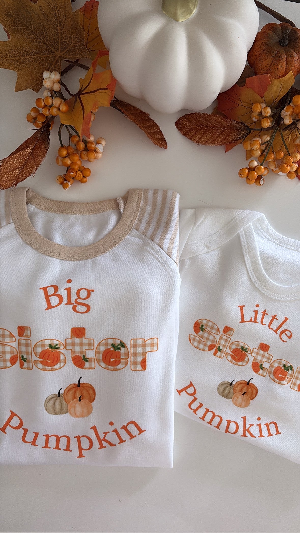 Two white baby t-shirts with beige ribbed trim, featuring autumn-themed 'Big Sister Pumpkin' and 'Little Brother Pumpkin' graphics, styled with fall leaves, berries, and a white pumpkin on a white background.