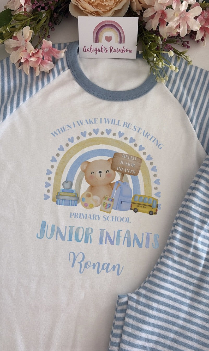 A white raglan-style baby/toddler shirt with a blue crew neck, laid flat on a blue-and-white striped fabric. The shirt has a colorful printed design featuring a teddy bear, rainbow, school books, a bus, and the text 'When I wake I will be starting Junior Infants Ronan.
