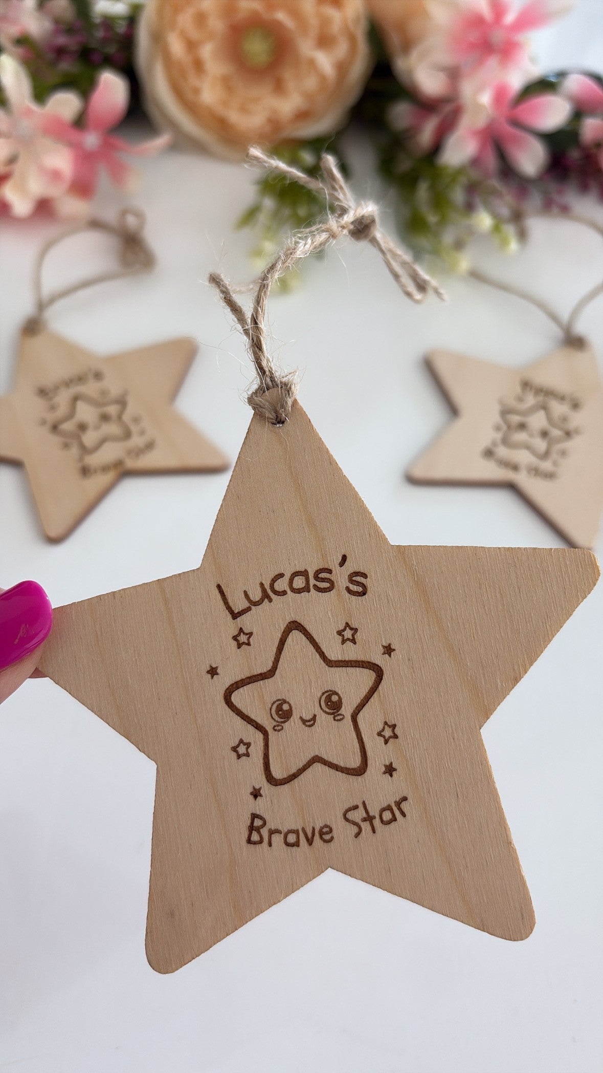 A wooden star-shaped hanging ornament engraved with 'Lucas's Brave Star' and a cute star face, held by a hand with pink nail polish. Two similar wooden stars are blurred in the background with flowers.