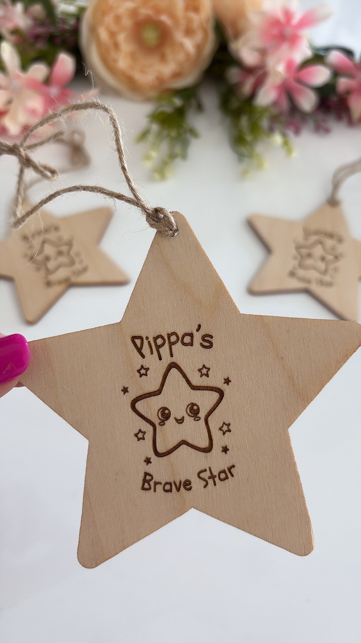 A wooden five-point star ornament engraved with 'Dippa's Brave Star' and a cute star face, held by a hand with pink nail polish. Other similar wooden ornaments are blurred in the background with decorative flowers.