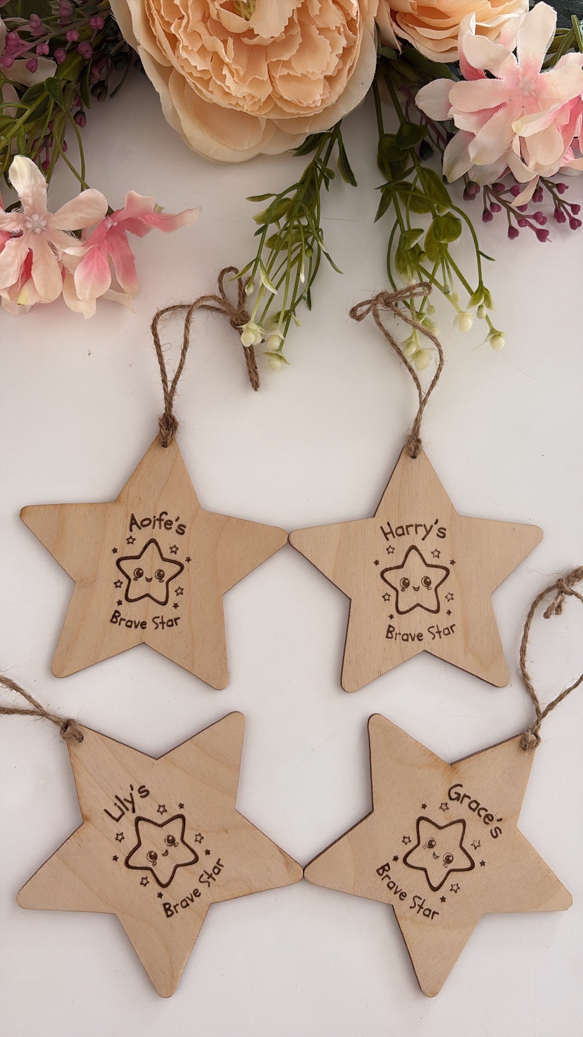 Four natural wood star-shaped hanging ornaments with engraved names and star illustrations, arranged on a white surface with decorative flowers around the top edge.
