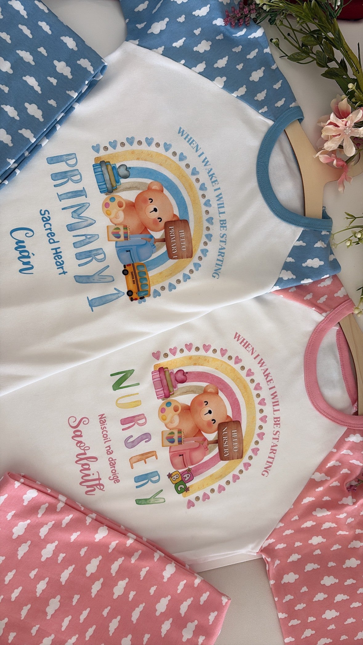 A styled flat-lay of baby bodysuits showing three color options: white with blue cloud-patterned sleeves and a cartoon bear graphic for 'Primary 1'; pink with white cloud-patterned sleeves and a cartoon bear graphic for 'Nursery'; and blue with white cloud-patterned sleeves and a cartoon bear graphic for 'Sweetheart'.