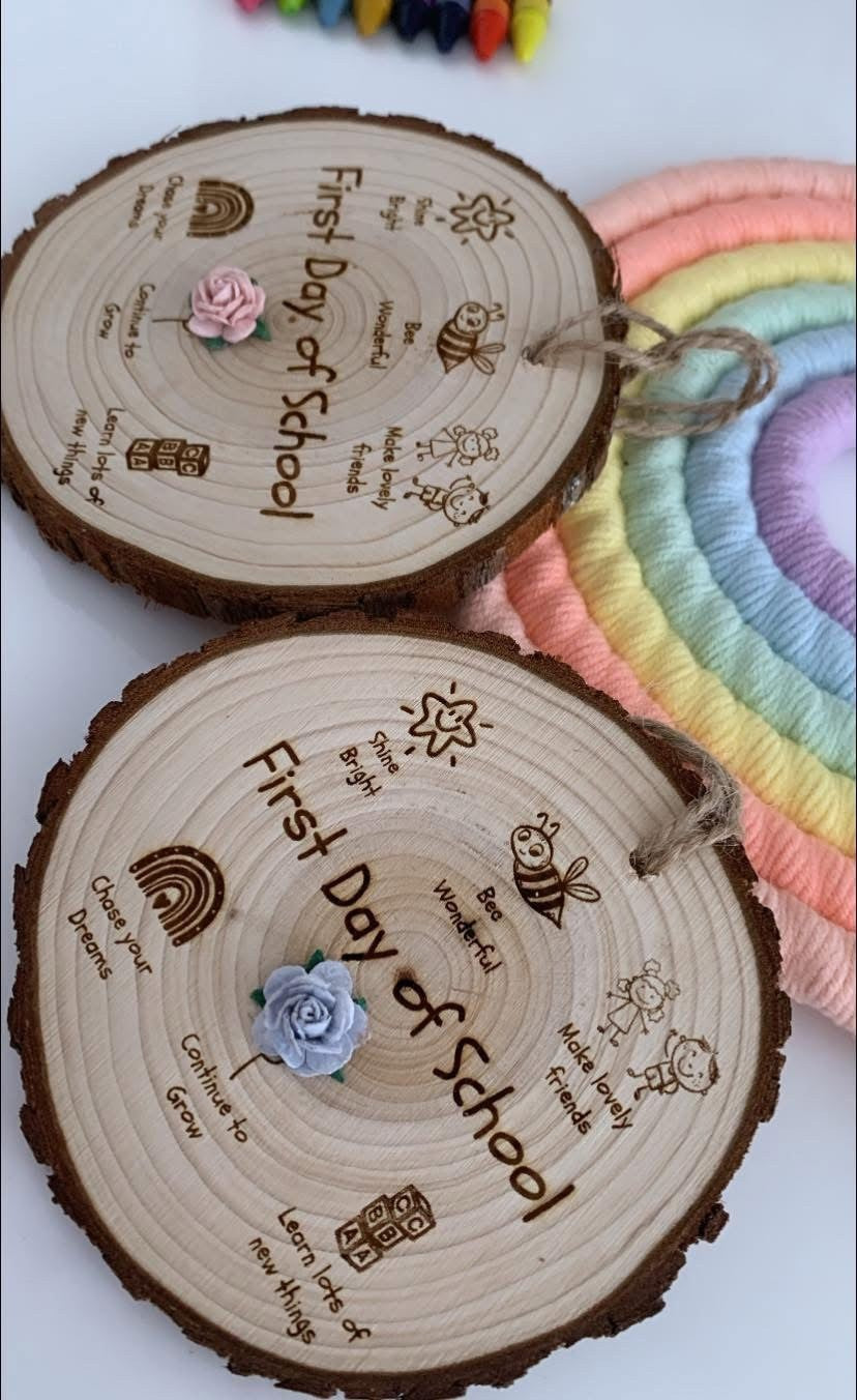 Two round engraved wooden tree slice plaques with 'First Day of School' text, hanging twigs, small fabric flowers, and a rainbow woven mat in the background.