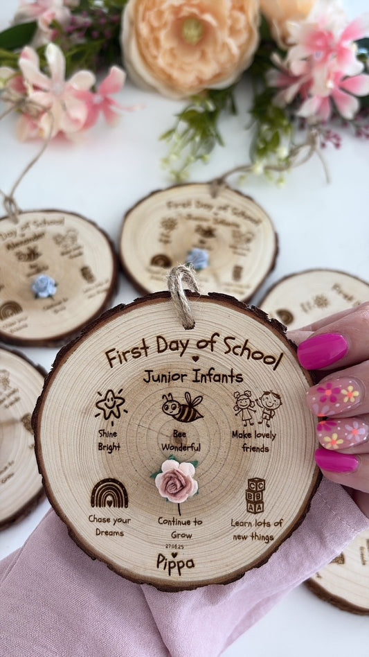 A personalized wooden slice keepsake tag for a child's first day of school, held by a hand with pink nail polish. The tag is engraved with 'First Day of School Junior Infants' and the name 'Pippa', decorated with a small pink rose.