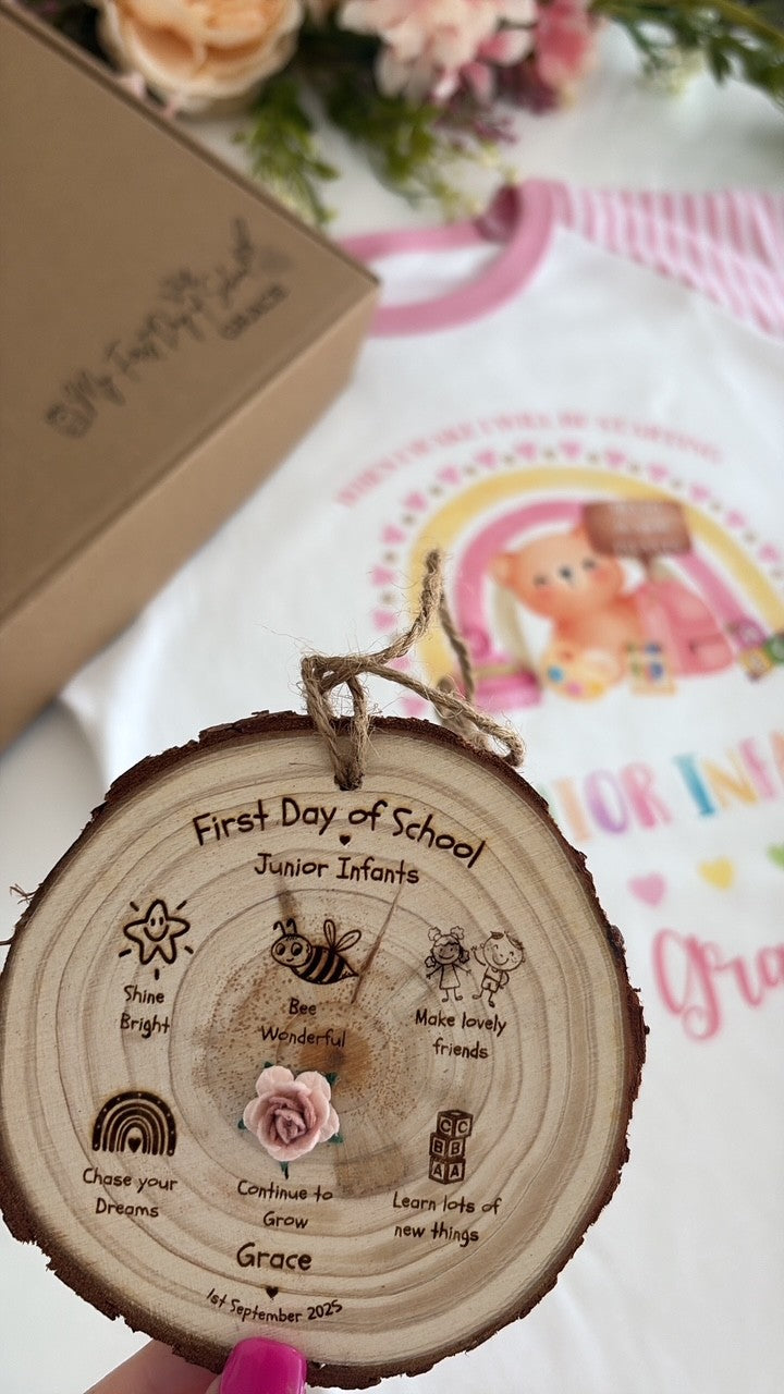 A personalized wooden log slice ornament engraved with 'First Day of School Junior Infants' and the name 'Grace', held by a hand. In the background, a white t-shirt with a pink and yellow cartoon bear graphic is visible next to a brown gift box.