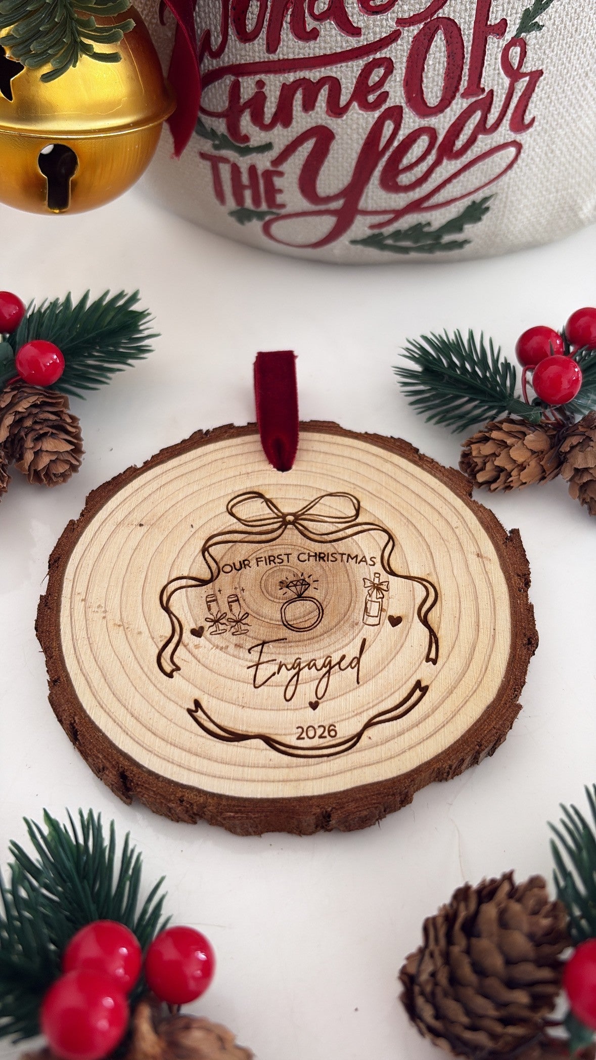 A personalized wooden Christmas ornament shaped like a tree slice, engraved with a ring and the words 'Engaged 2026' and 'Your First Christmas'. The ornament hangs from a red ribbon and is styled on a white surface with pinecones, red berries, green pine branches, and a gold bell in the background.