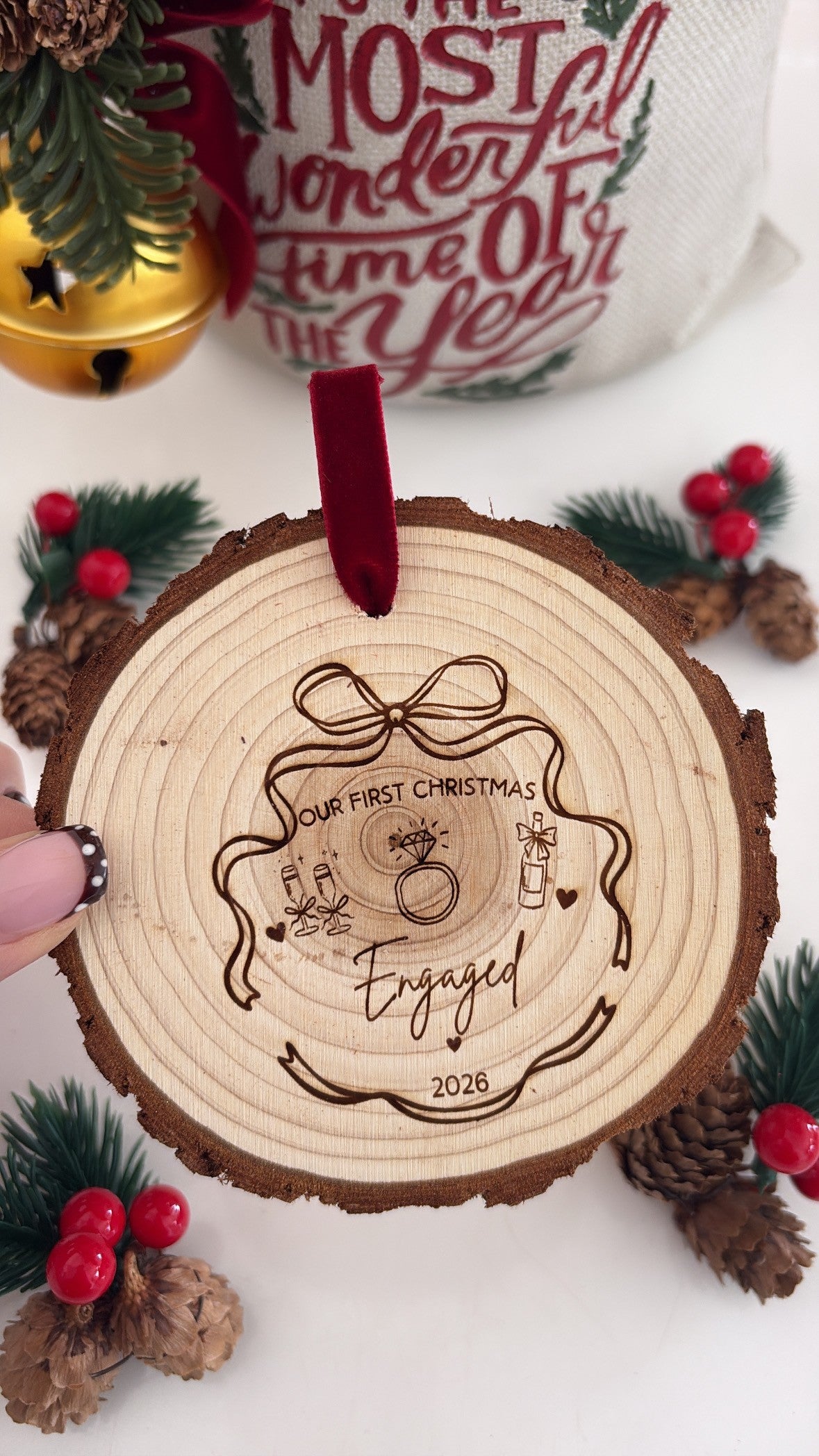 A personalized wooden Christmas ornament featuring a laser-engraved design of a Christmas tree and the words 'Our First Christmas Engaged 2026' with a red ribbon loop, held by a hand against a festive holiday background.