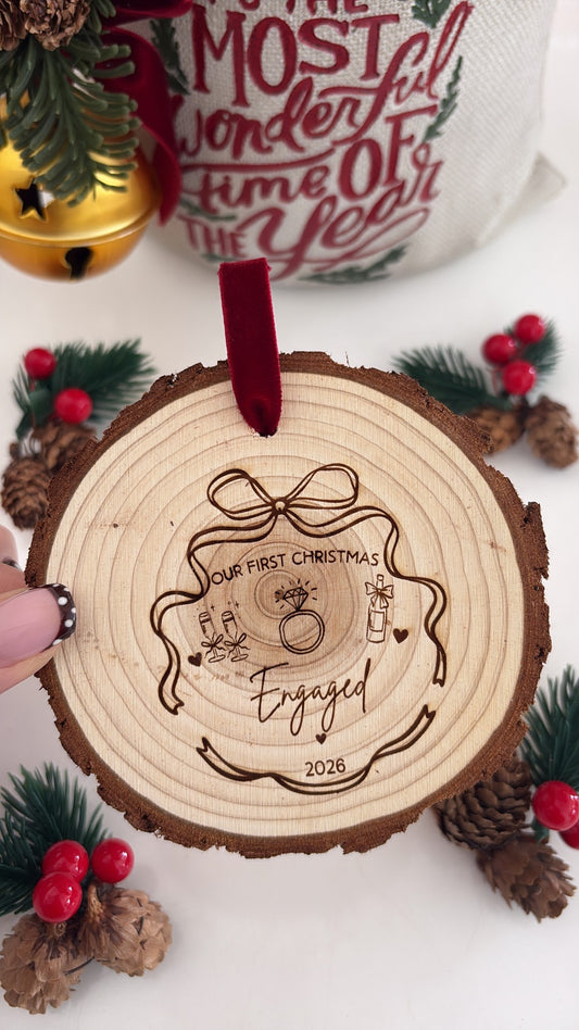 A personalized wooden Christmas ornament featuring a laser-engraved design of a Christmas tree and the words 'Our First Christmas Engaged 2026' with a red ribbon loop, held by a hand against a festive holiday background.