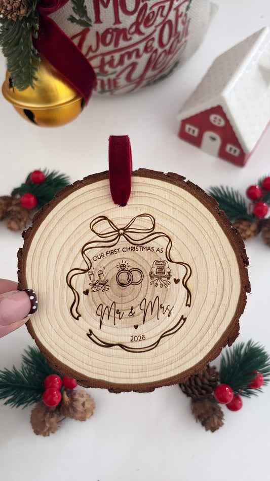 A round wooden Christmas ornament with a natural wood slice edge, featuring a red hanging loop and an engraved design that reads 'Our First Christmas As' and 'Mr & Mrs 2026' with a ring motif. A hand is holding the ornament, and the background includes festive holiday decorations like pine branches, red berries, a small red house, and a gold bell.