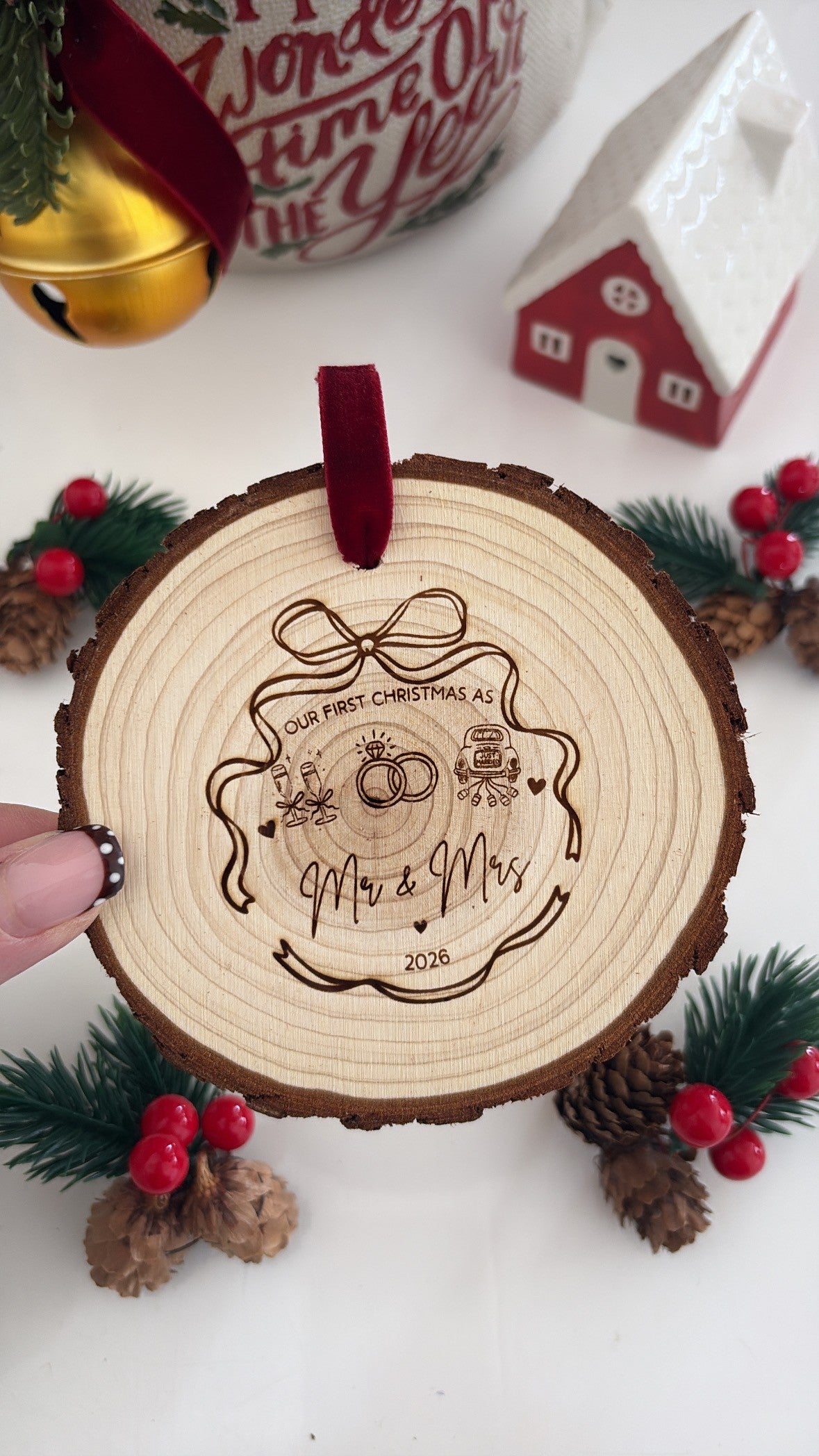 A personalized wooden Christmas ornament shaped like a tree bark slice, held by a hand. The wood is engraved with 'Our First Christmas As Mr & Mrs 2025' with a ring and gift box illustration, and features a dark red velvet hanging loop.