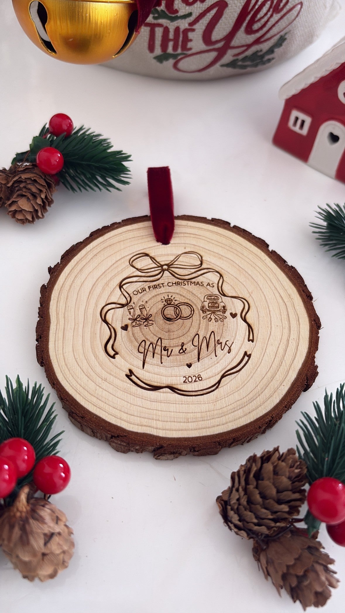 A personalized wooden Christmas ornament featuring a laser-engraved design of two wedding rings and the text 'Mr & Mrs 2026', hanging from a dark red ribbon. The ornament is styled on a white surface with pinecones, green pine branches with red berries, and a festive background.
