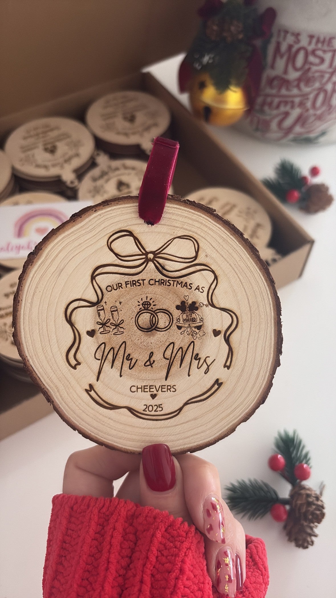 A personalized wooden Christmas ornament held by a hand, featuring an engraved design for a first Christmas together with a red velvet ribbon loop. The background shows a box of similar wooden ornaments and festive decorations.