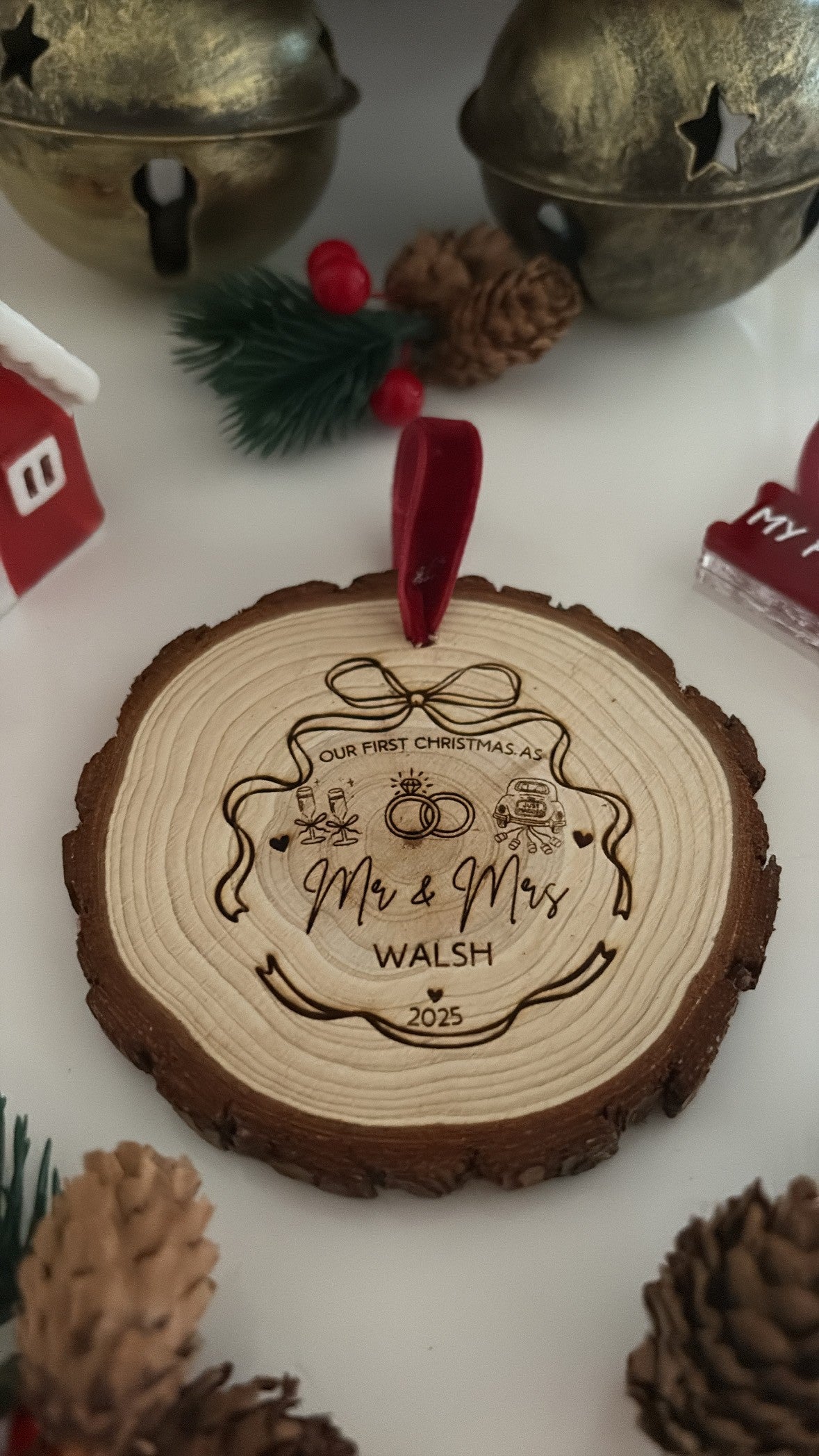 A personalized Christmas ornament made from a natural wood slice with a red hanging loop, featuring engraved text and illustrations including 'Our First Christmas As Mr & Mrs Walsh 2025'. The ornament is styled on a white surface with pinecones, artificial greenery, and brass bells in the background.