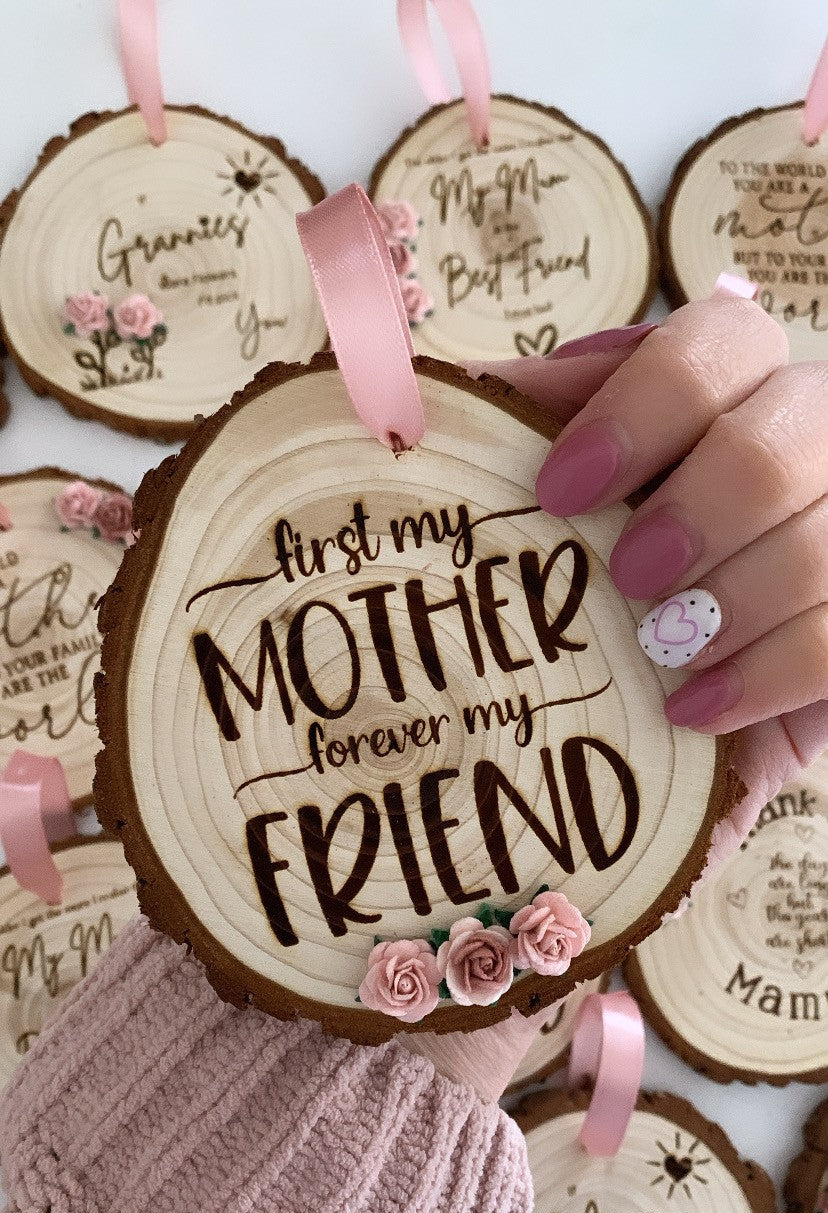 Hand holding a wooden hanging disc engraved with “First my mother, forever my friend”, decorated with pink flowers and a ribbon, surrounded by similar wooden keepsakes.