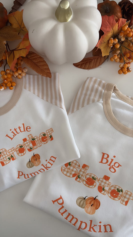 Two white baby t-shirts with beige trim, personalized with 'Little Brother Pumpkin' and 'Big Sister Pumpkin' in orange text with a plaid 'O' and small pumpkin illustrations. The shirts are styled in a flat-lay with fall decorations including a white pumpkin, autumn leaves, and berries.