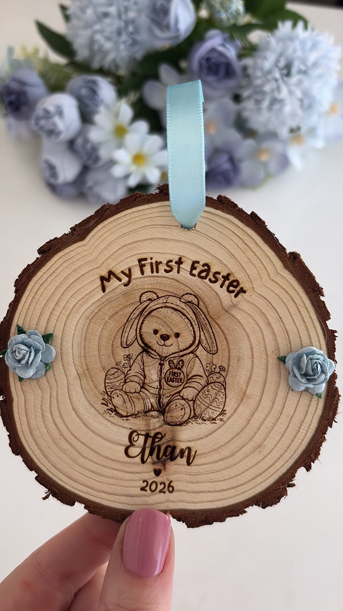 Personalised “My First Easter” wooden disc with engraved bunny teddy design, child’s name and year, rustic bark edge and blue ribbon hanger.