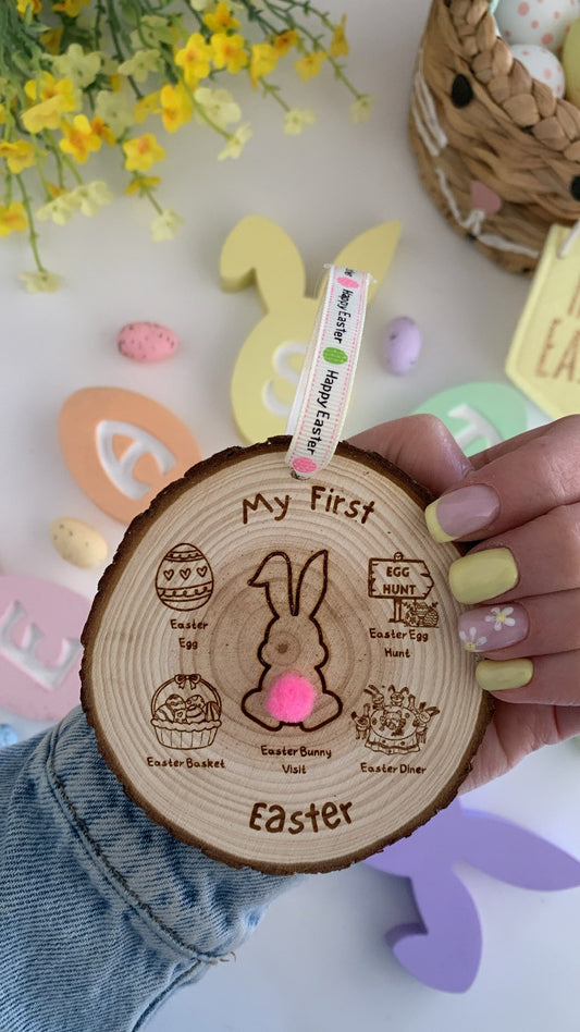 Wooden “My First Easter” milestone disc with engraved bunny design, blue pom-pom tail and Easter themed illustrations, hanging with pastel ribbon.