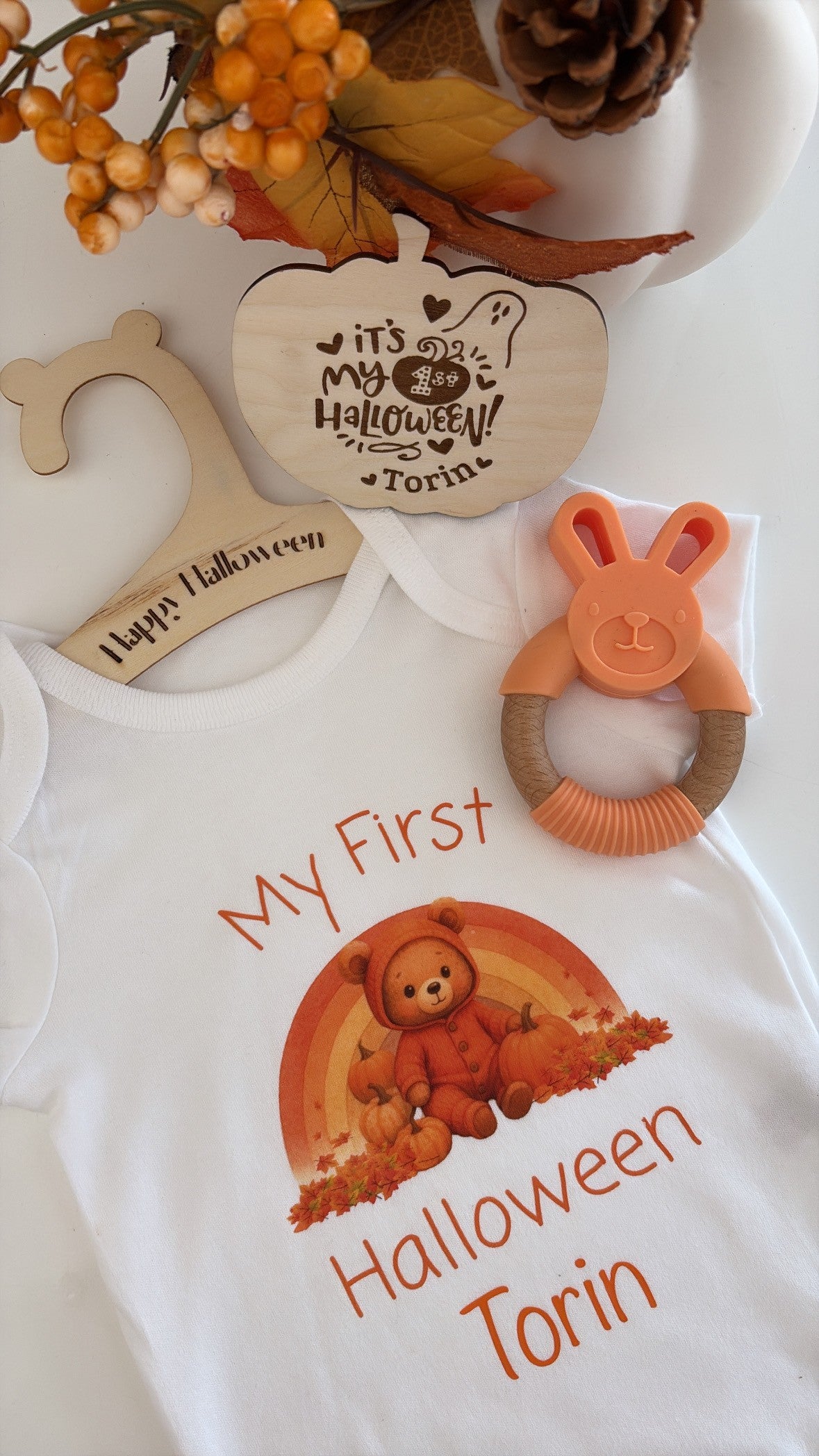 A white baby bodysuit with a custom printed graphic reading 'My First Halloween Torin' in orange text with a teddy bear and pumpkins, styled with wooden hangers, a bunny teether, and autumn decorations.