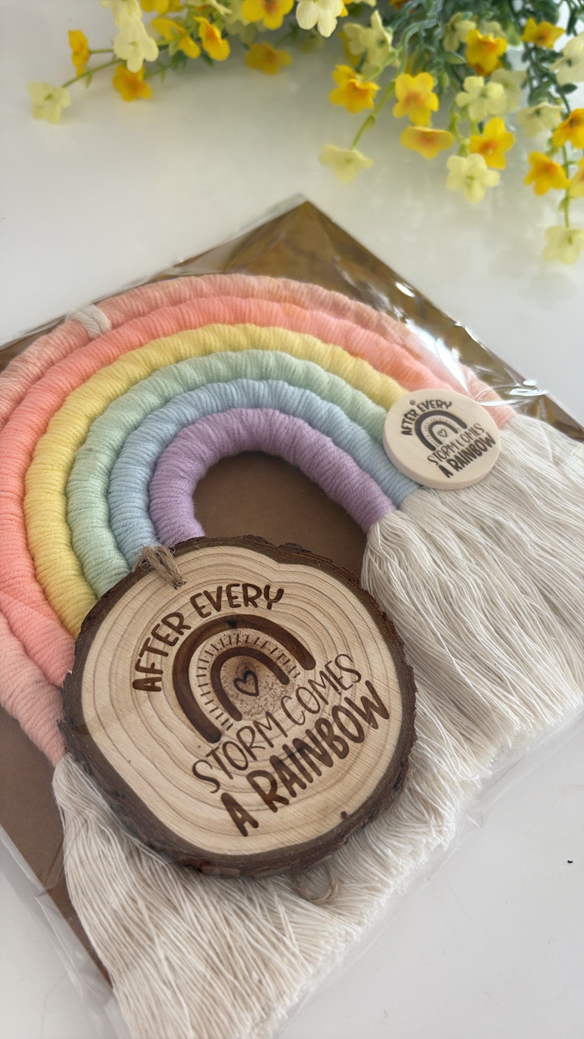 Wooden round tag with 'After every storm comes a rainbow' text, surrounded by colorful yarn and flowers.