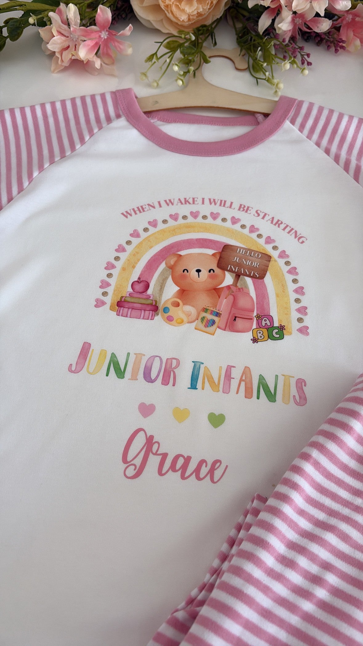 A white raglan baby t-shirt with pink striped sleeves and a pink crew neck, featuring a cute cartoon teddy bear and rainbow graphic with the text 'JUNIOR INFANTS Grace'. The shirt is shown on a wooden hanger with pink striped fabric and decorative flowers in the background.