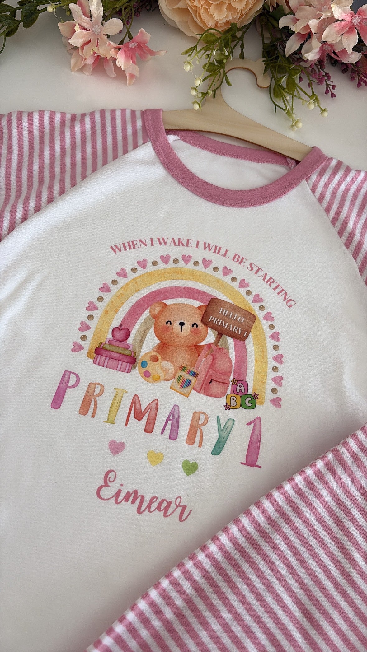 A white raglan-style t-shirt with pink trim and sleeves, featuring a colorful 'Primary 1' birthday graphic with a bear, rainbow, and cake. The shirt is displayed on a wooden hanger over a pink and white striped blanket with decorative flowers in the background.