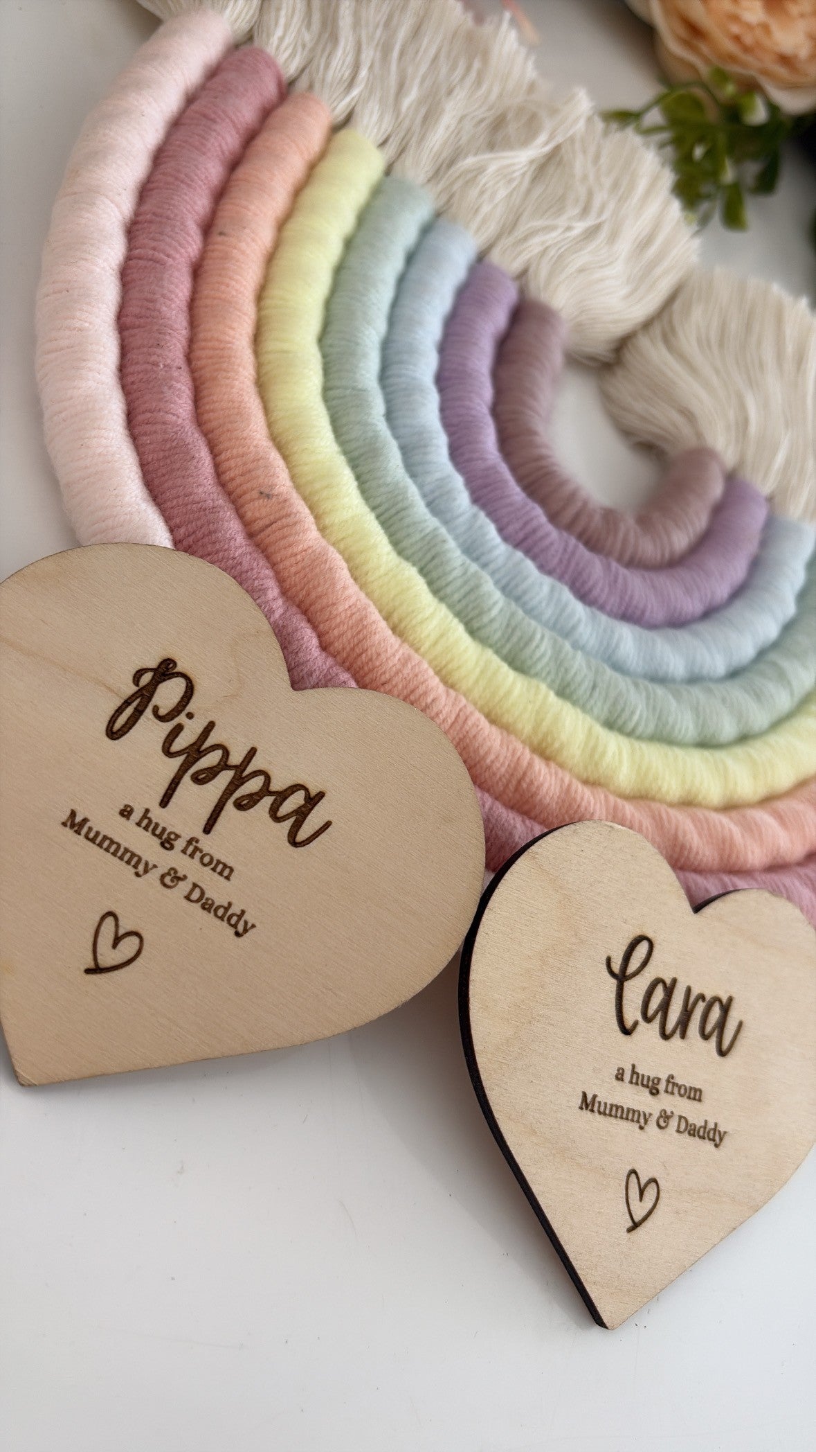 Two personalized wooden heart-shaped plaques engraved with names and a message from mum and dad, placed in front of a pastel rainbow macrame wall hanging on a white surface.