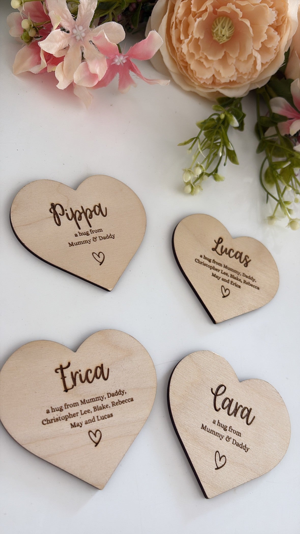 Four personalized heart-shaped wooden place cards with engraved names and messages, arranged on a white surface with decorative flowers in the corners.