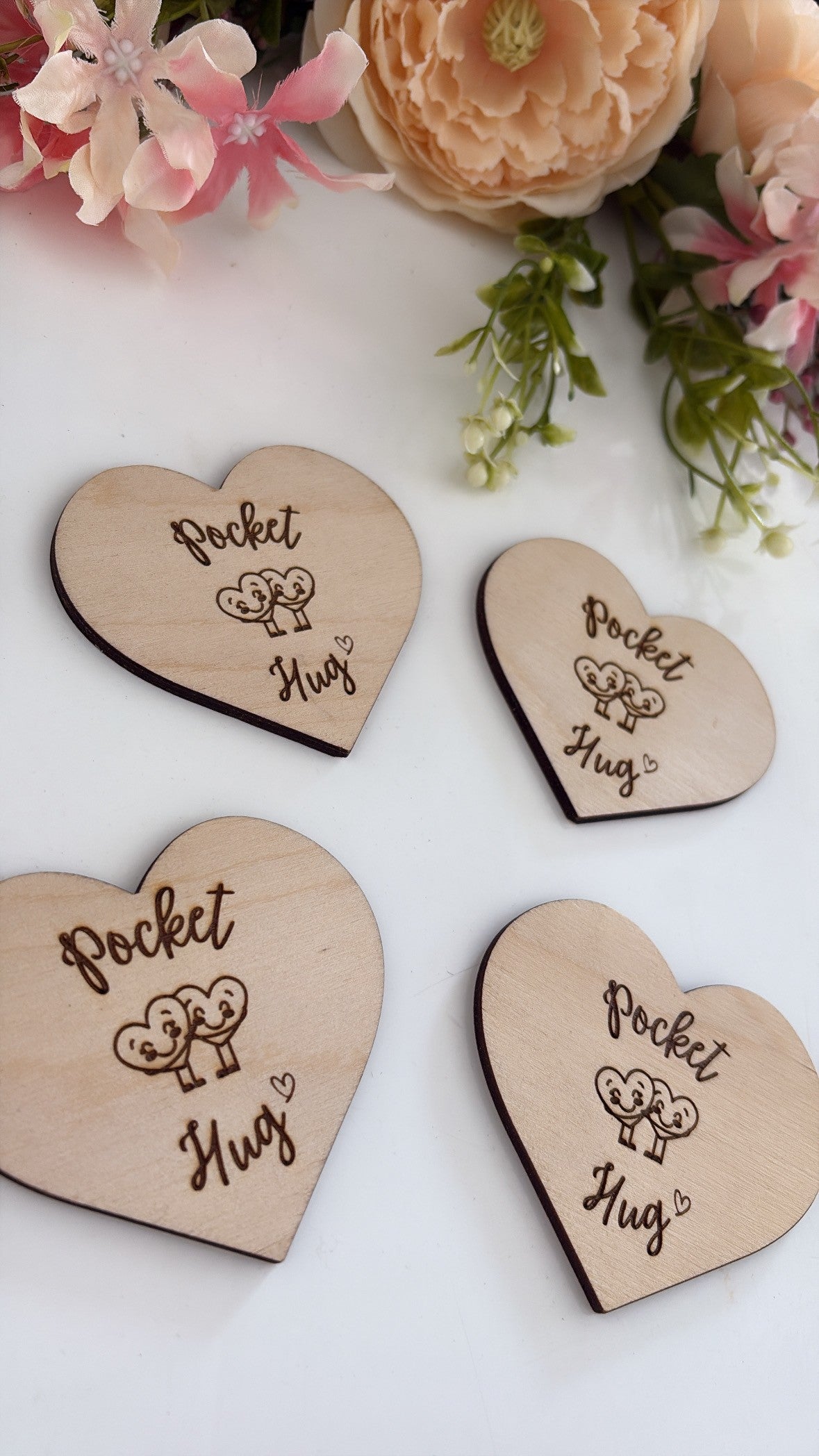 Four heart-shaped wooden coasters engraved with 'Pocket Hug' and a small heart design, arranged on a white surface with decorative pink flowers in the background.