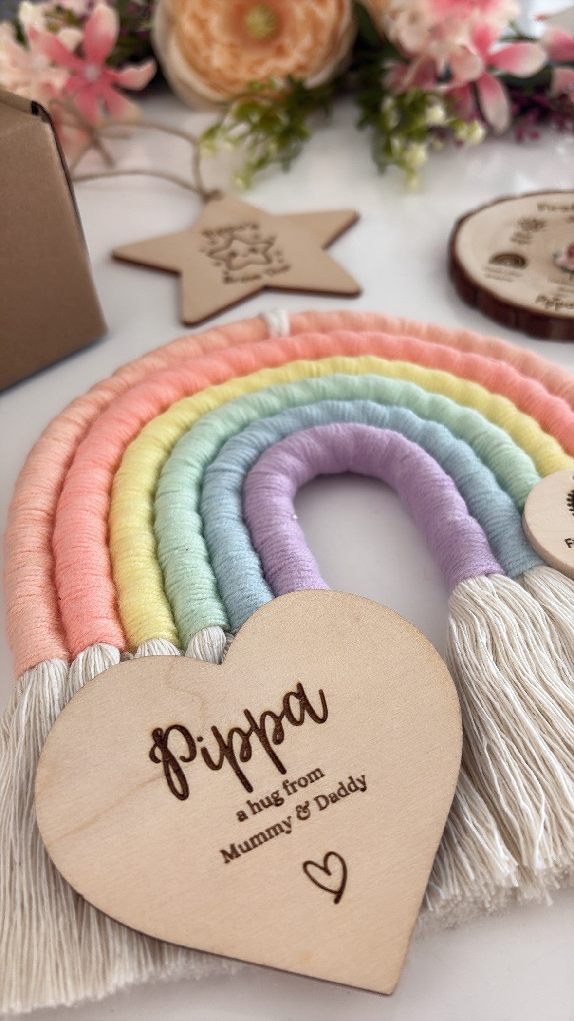 A pastel rainbow macrame wall hanging with white tassels and a personalized wooden heart plaque that reads 'Pippa a hug from Mummy & Daddy'. The photo is styled with flowers and other wooden decor on a white surface.