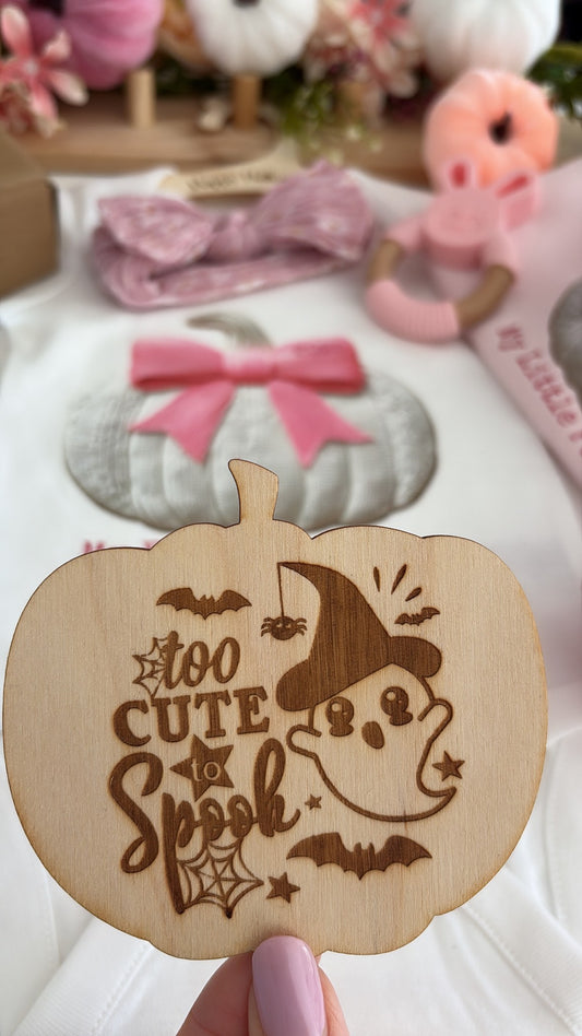 A hand with pink nail polish holds a wooden pumpkin-shaped sign engraved with 'too CUTE Spook' and a cartoon ghost in a witch hat. The background shows a styled flat-lay of baby accessories, including a pink bow headband, a gray pumpkin patch, and a pink pacifier clip.