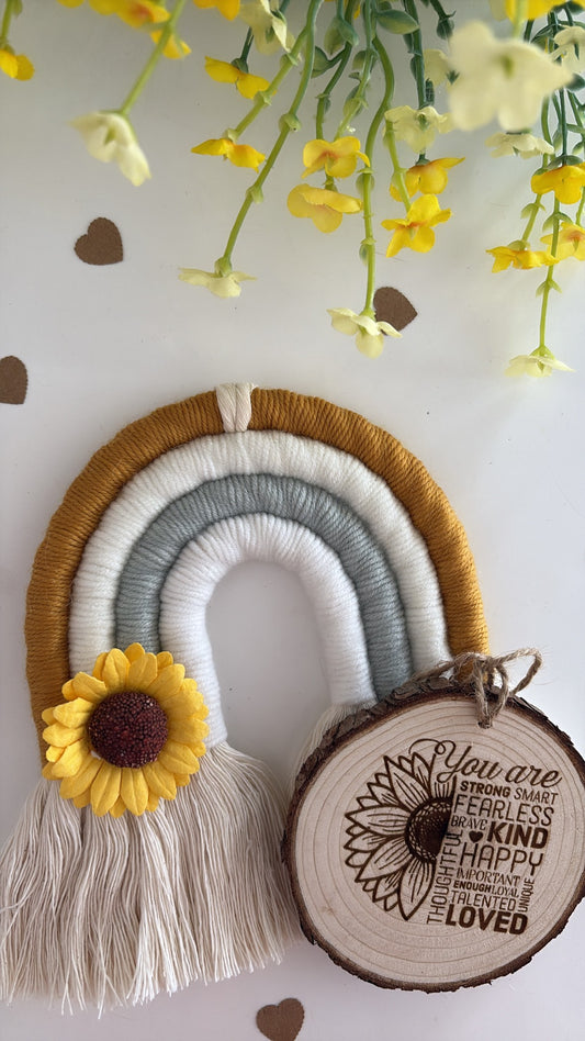 “Handmade macrame rainbow in mustard, cream, and gray with a felt sunflower and engraved wooden tag that reads positive affirmations, displayed with yellow flowers