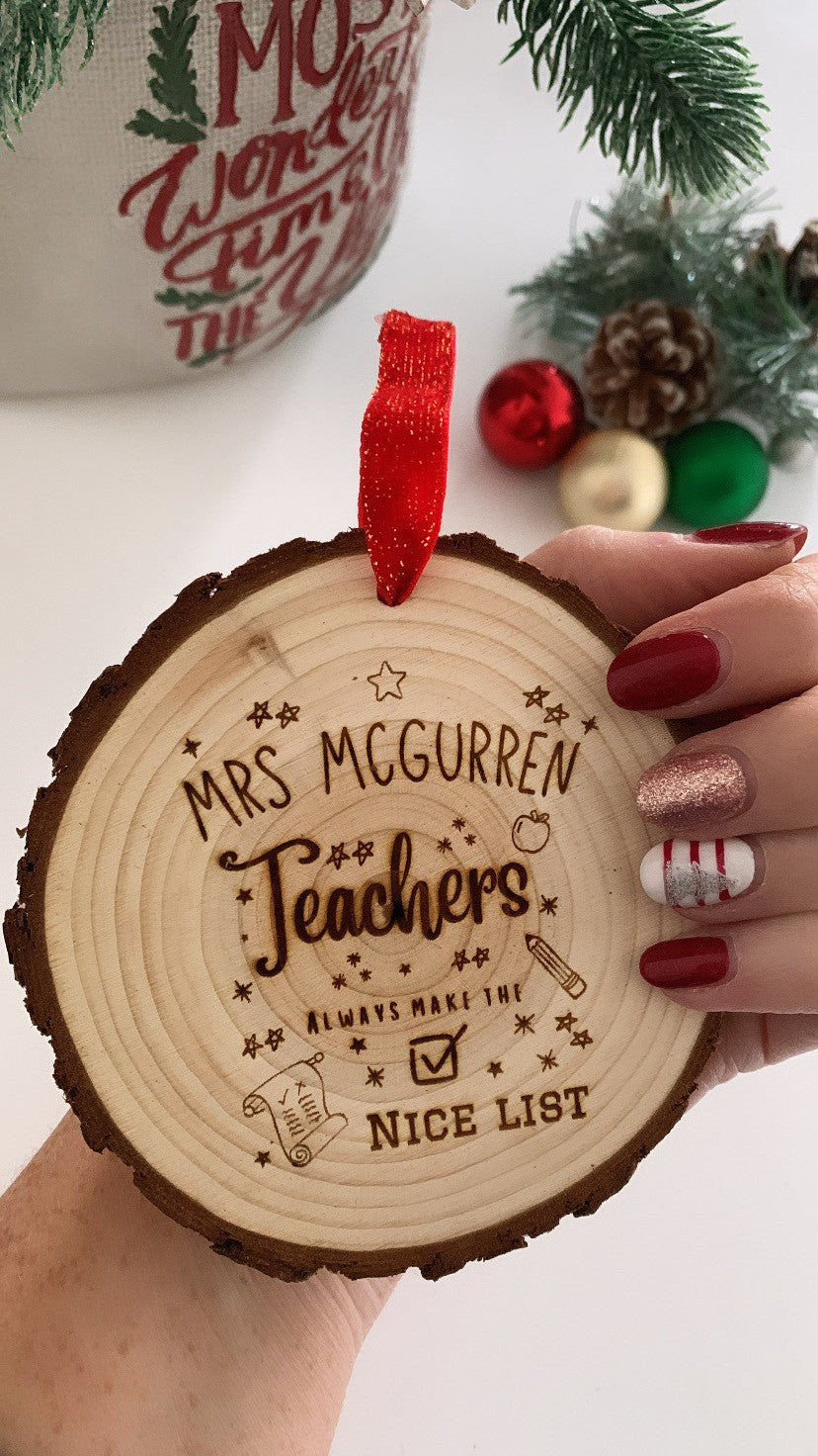 Wooden ornament with engraved text held by a hand, with Christmas decorations in the background. Teachers always make the nice list